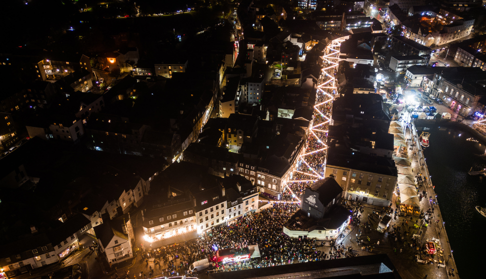 Aerial night view of a bustling city street decorated with zigzagging strings of lights. Thousands of people are gathered along the brightly lit street and outside surrounding buildings, creating a festive atmosphere.